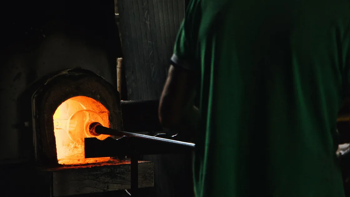Vidriero de espaldas soplando una pieza frente a la boca encendida del horno de un taller